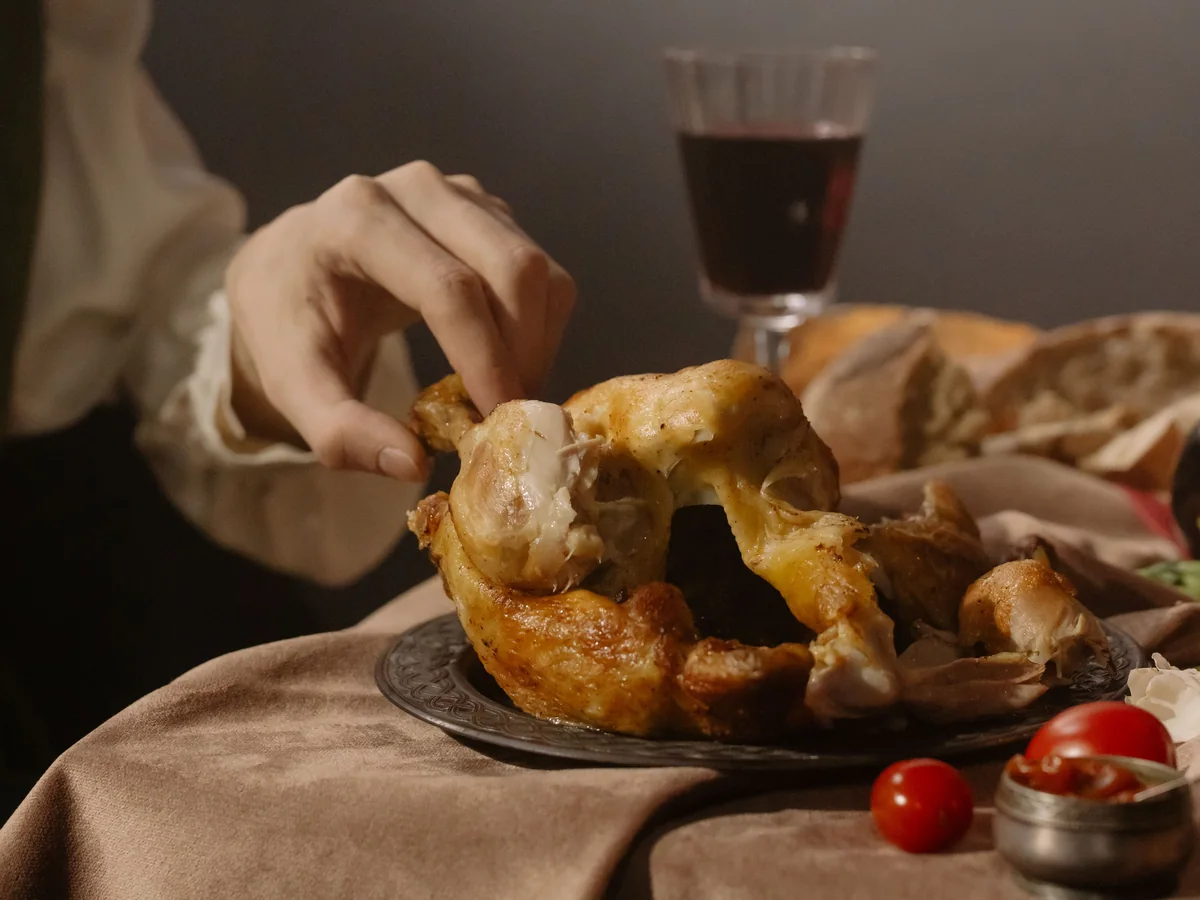 A hand pulling apart a whole roast chicken by hand, with wine and bread on the table