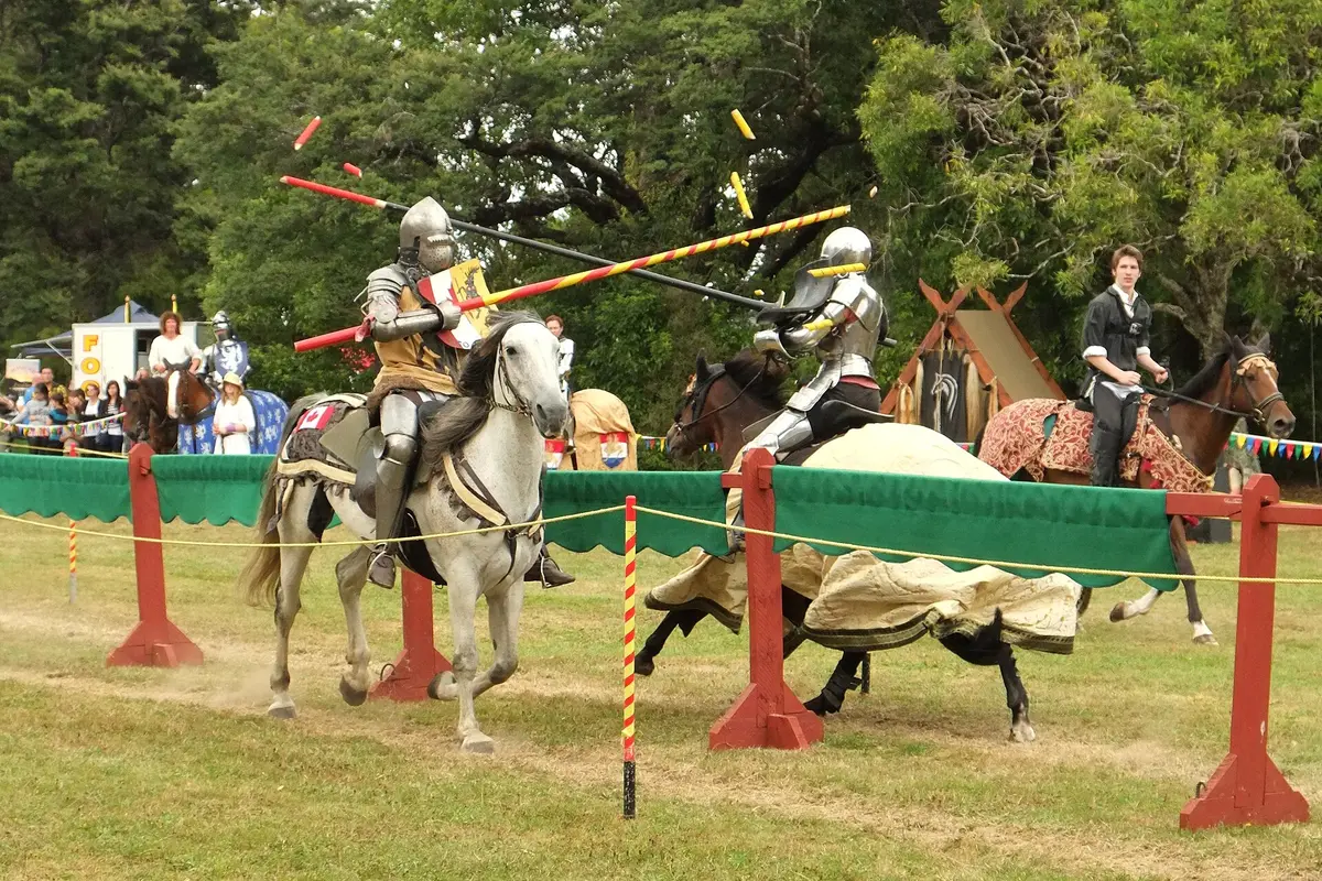 Two armoured knights on horseback pass each other at speed, their lance tips splintering into the air, behind a low barrier on a grass field