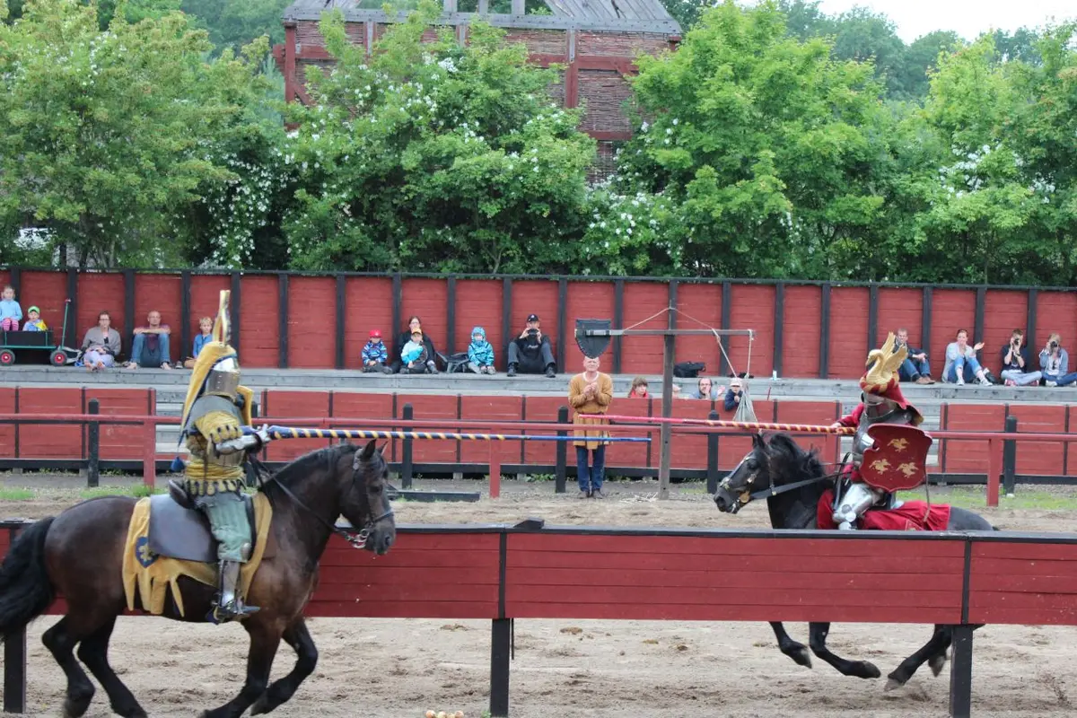 Two knights on horseback charge towards each other along a red wooden barrier in a sand arena, with spectators seated behind a fence